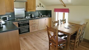 A kitchen with wooden cabinets, a dining table with chairs, and a window with a red valance. - 2 bedroom accommodation in Pendeen (Pendeen)
