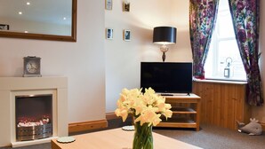 Een woonkamer met een open haard, een flatscreen televisie, een tafel met een bloemenvaas en een raam met gordijnen.