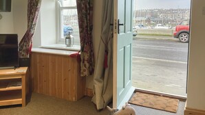 Een kamer met een houten bankje, een raam met bloemen gordijnen, en uitzicht op een straat met geparkeerde auto's en gebouwen.