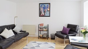 A modern living room with a black leather sofa, a black armchair, and a small table with a vase of flowers. There is a radiator, a bookshelf, and a geometric rug on the floor. - 2 bedroom accommodation in Bonar Bridge (Bonar Bridge)
