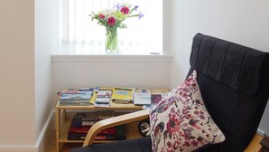 A room with a black armchair, a wooden side table with books and a vase of flowers, and a window with blinds. - 2 bedroom accommodation in Bonar Bridge (Bonar Bridge)