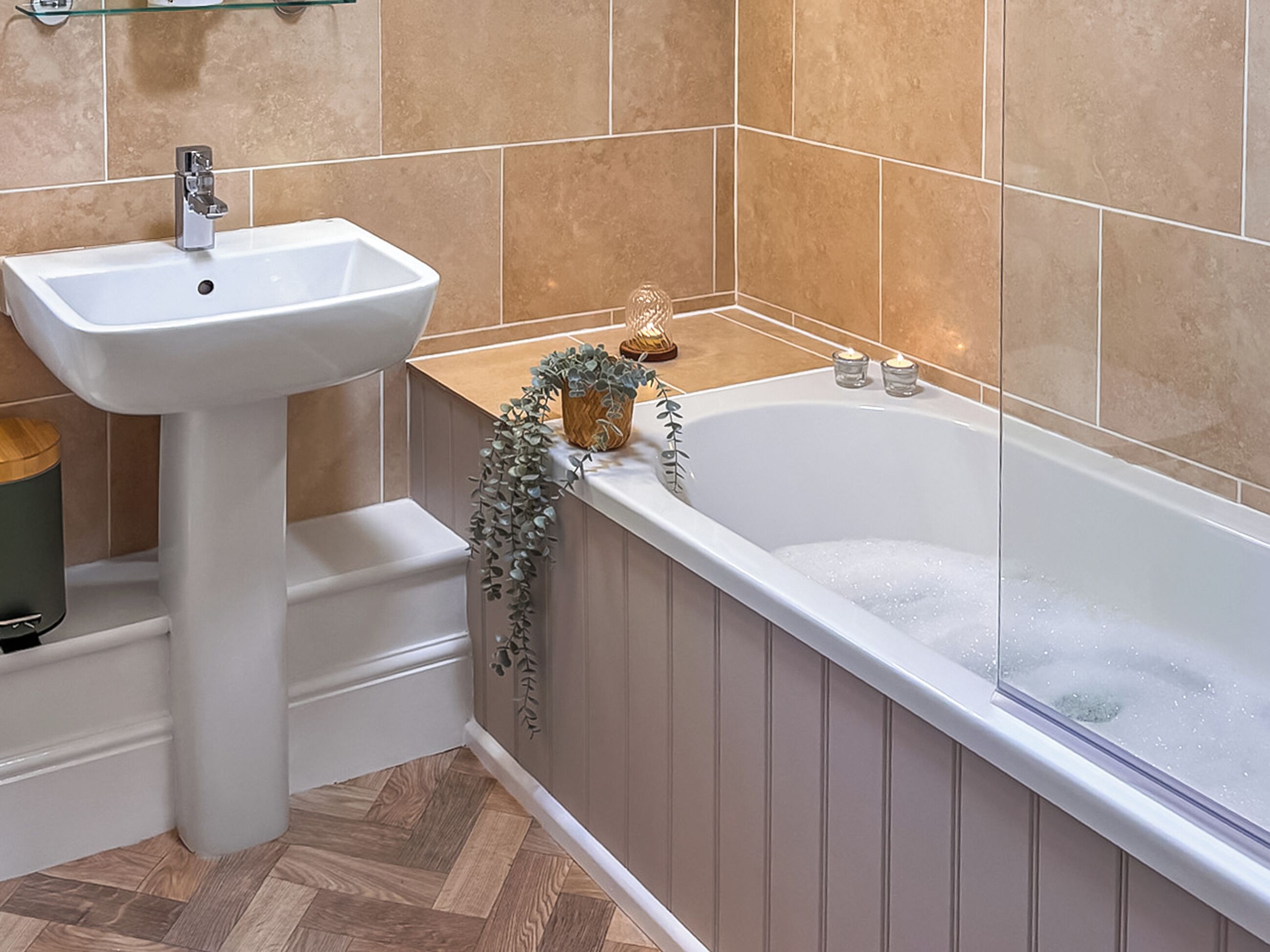 A bathroom with a white pedestal sink, a bathtub with bubbles, and a wooden floor.