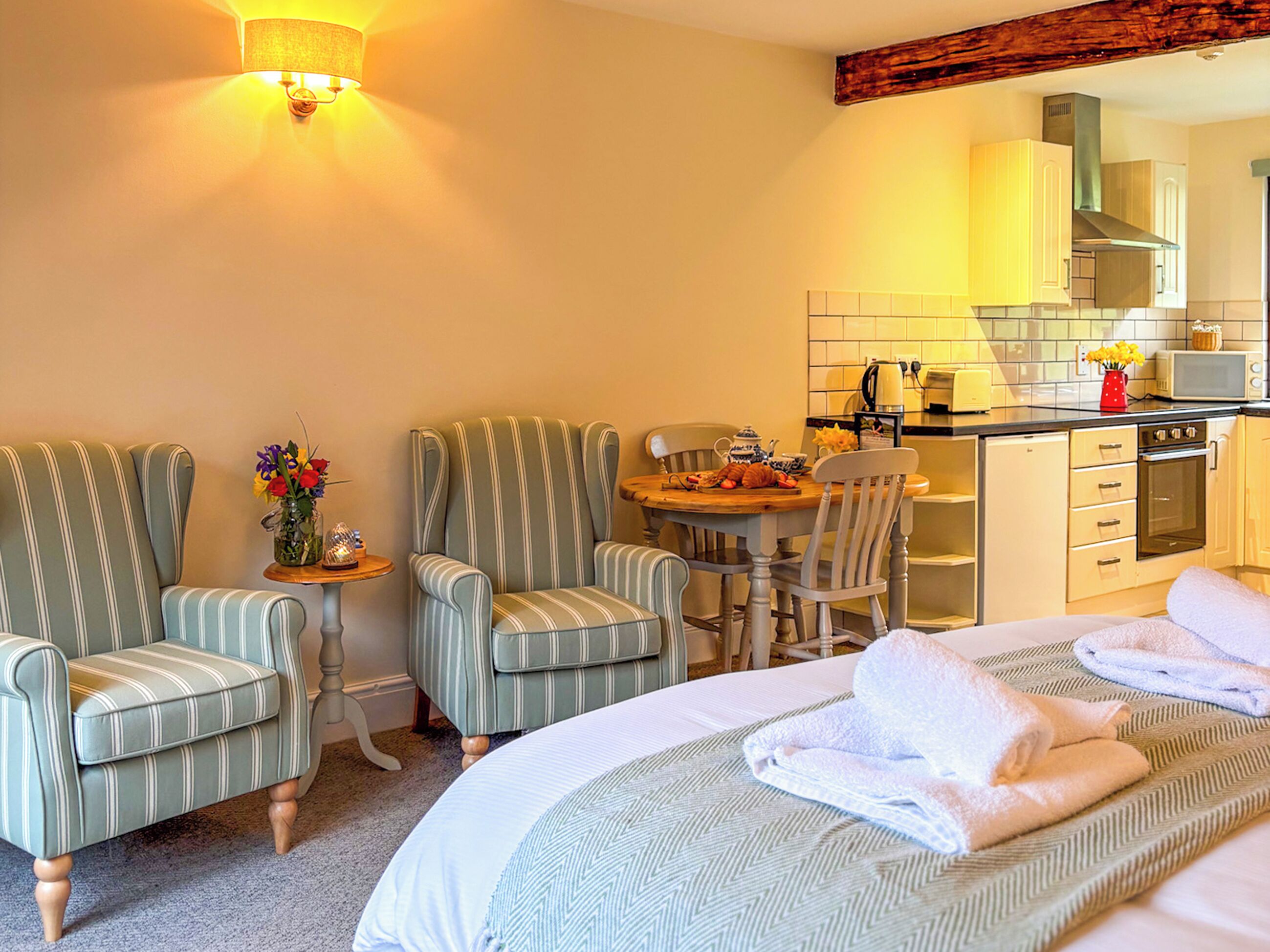 A hotel room with a bed, two striped armchairs, a small table with a vase of flowers, a kitchen area with a microwave, and a stove.