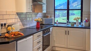Une cuisine avec des meubles blancs, un sol à motifs en chevrons, un grille-pain, un four à micro-ondes et une fenêtre donnant sur un paysage verdoyant.