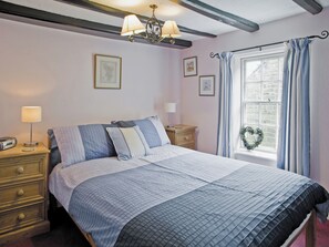 A bedroom with a bed, bedside tables, a dresser, a window with curtains, and a chandelier. - Sharrowhead Cottage - E4044 (Robin Hood’s Bay)