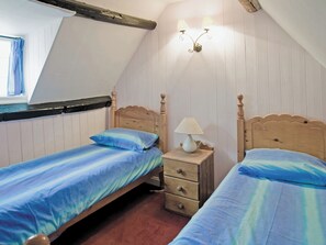 A bedroom with two single beds, a nightstand with a lamp, a window with curtains, and wooden paneling on the walls. - Sharrowhead Cottage - E4044 (Robin Hood’s Bay)