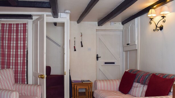 A room with two striped sofas, a small wooden table, a door with a red curtain, and a wall-mounted light fixture. - Sharrowhead Cottage - E4044 (Robin Hood&rsquo;s Bay)