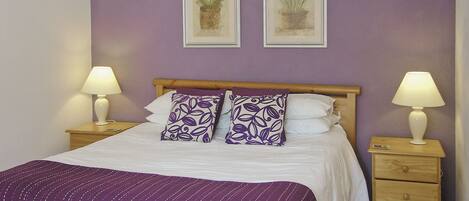 Una cama bien hecha con un edredón morado, dos lámparas de noche y dos cuadros en la pared.