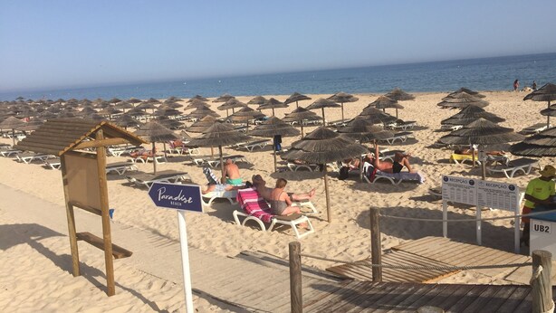 A beach with numerous sun umbrellas and lounge chairs, a wooden boardwalk, and a sign indicating 'Paradise Beach'.
