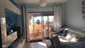 A modern living room with a sofa, a TV, and a balcony with a view of palm trees and buildings.
