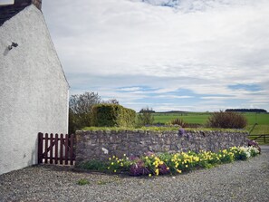 Un edificio rural con un muro de piedra y un camino de grava que conduce a un portón de madera.