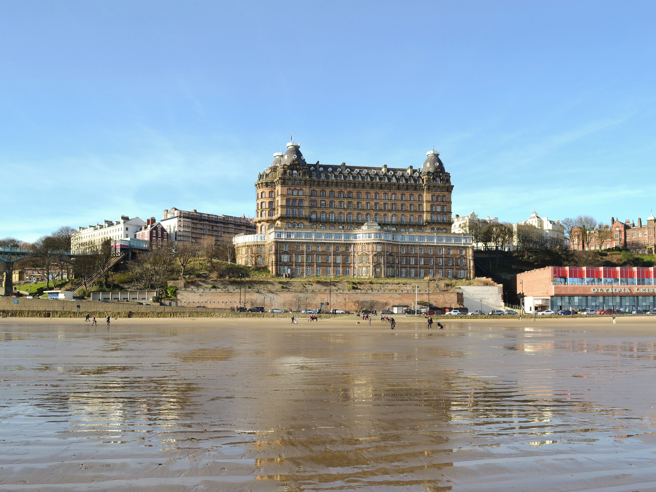 A large, historic building with multiple stories and ornate architecture, situated by a waterfront with a sandy beach and a calm body of water.