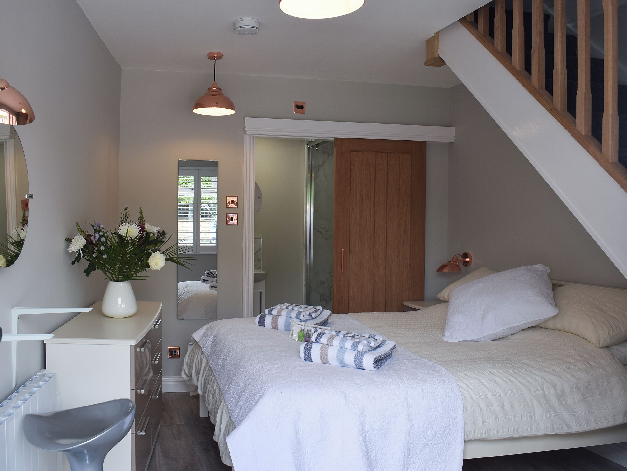 A bedroom with a bed, bedside table, and a desk with a chair. There is a mirror on the wall, a window with blinds, and a wooden door.