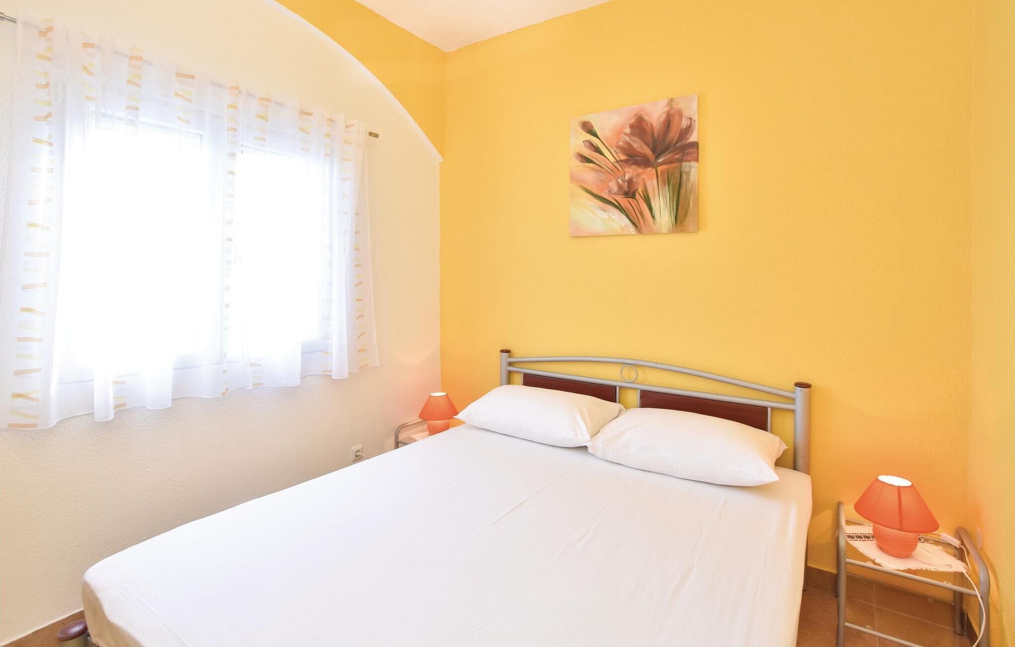 A bed with white linens in a room with yellow walls, a window with sheer curtains, and a painting of flowers on the wall.