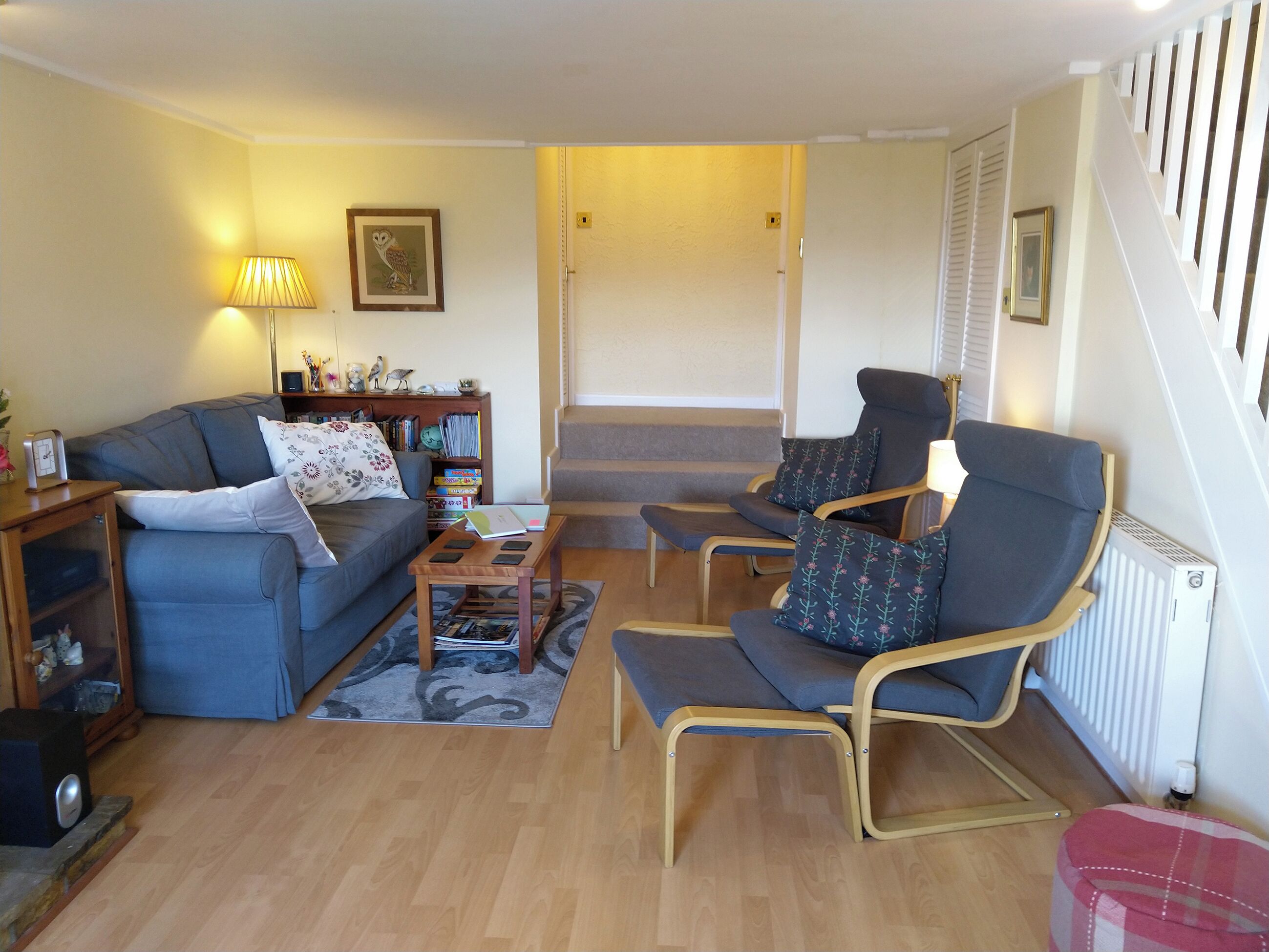 A living room with a sofa, a chair, a coffee table, and a bookshelf. There is a staircase in the background.