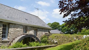 Un bâtiment en pierre doté d’un toit en pente, d’une arche en pierre et d’une cour verdoyante bordée d’un mur en pierre.