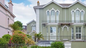 A large, two-story house with a balcony, surrounded by a stone wall and lush greenery. - 2 bedroom accommodation in Brixham (Brixham)