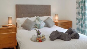 A neatly made bed with pillows, a bedside table with a lamp, a tray with breakfast items, and a window with patterned curtains. - 2 bedroom accommodation in Brixham (Brixham)