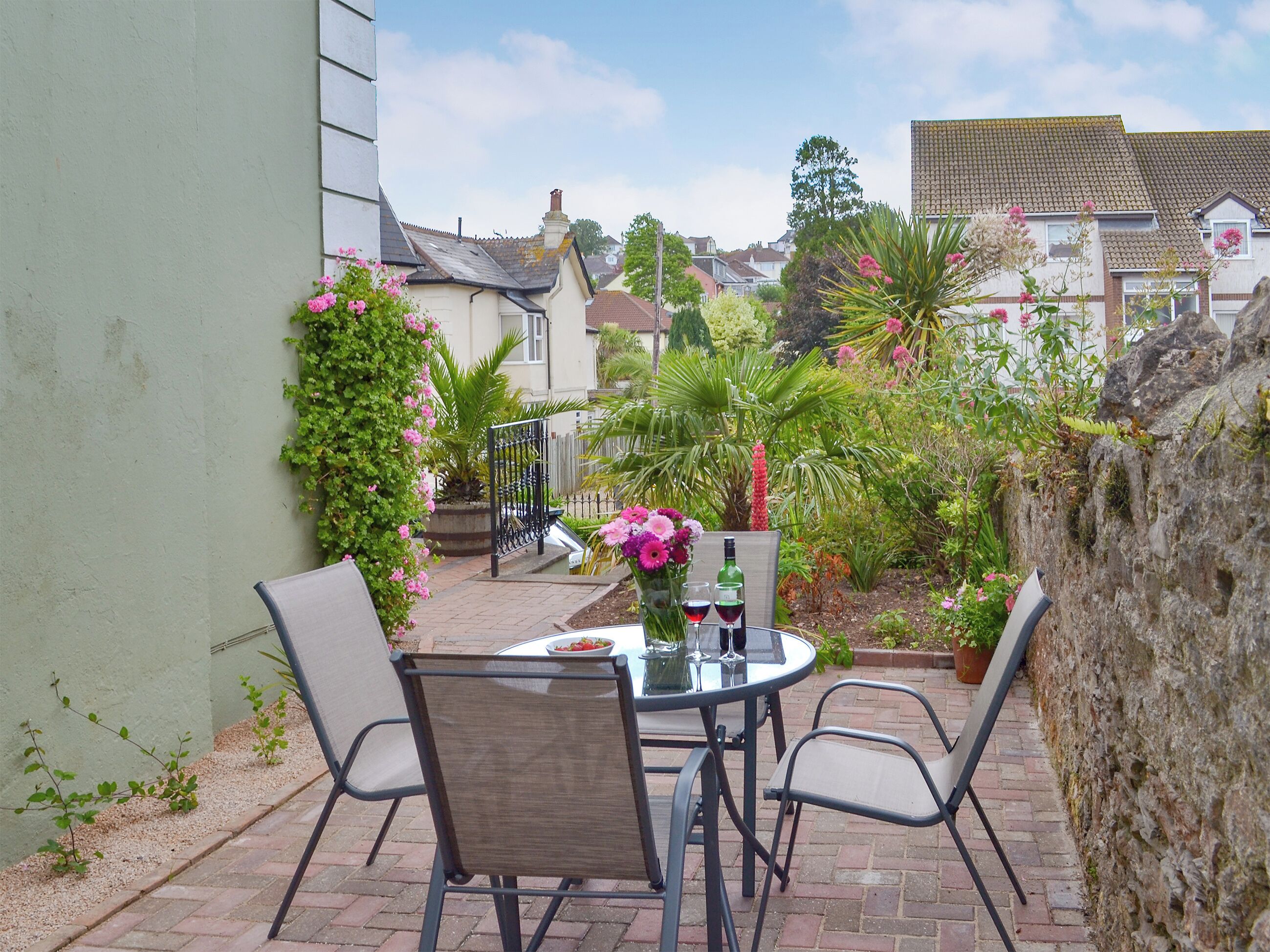 An outdoor patio with a table and chairs, surrounded by greenery and a view of houses in the background.