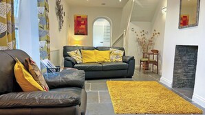 A modern living room with a black leather sofa, yellow cushions, and a yellow rug. There is a fireplace, a staircase, and a view of the hallway. - 2 bedroom accommodation in Brixham (Brixham)