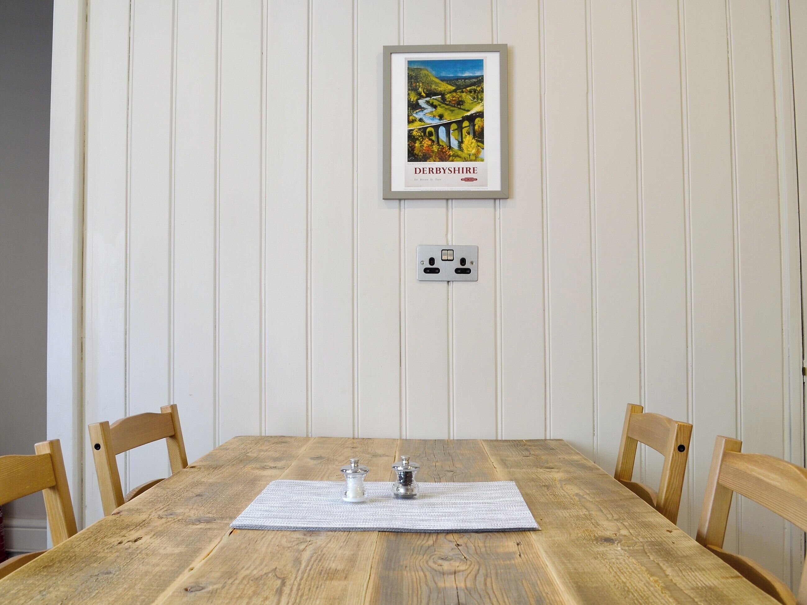 A wooden dining table with a placemat, two salt and pepper shakers, and a framed picture of a bridge in Derbyshire.