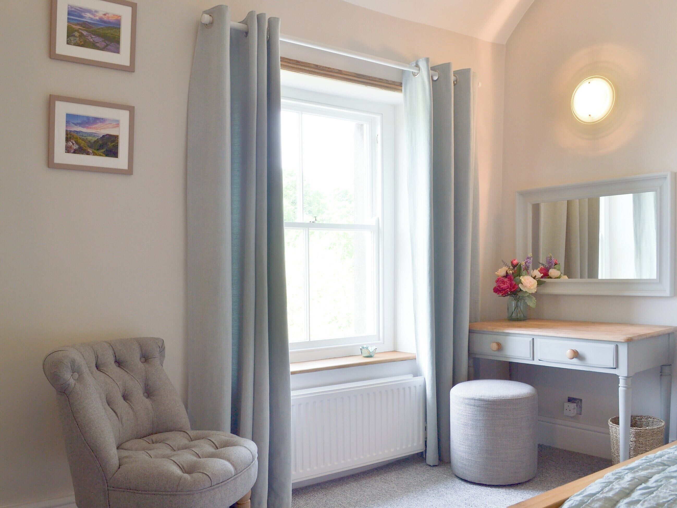 A bedroom with a window, a chair, a vanity with a mirror, and a bedside table.