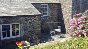Un bâtiment en pierre avec une fenêtre et une porte, un petit patio avec une table et des chaises, et un parterre de fleurs avec des fleurs roses.