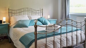 A neatly made bed with a metal frame, a bedside table with a lamp, and a window with curtains. - 4 bedroom accommodation in Keswick (Keswick)