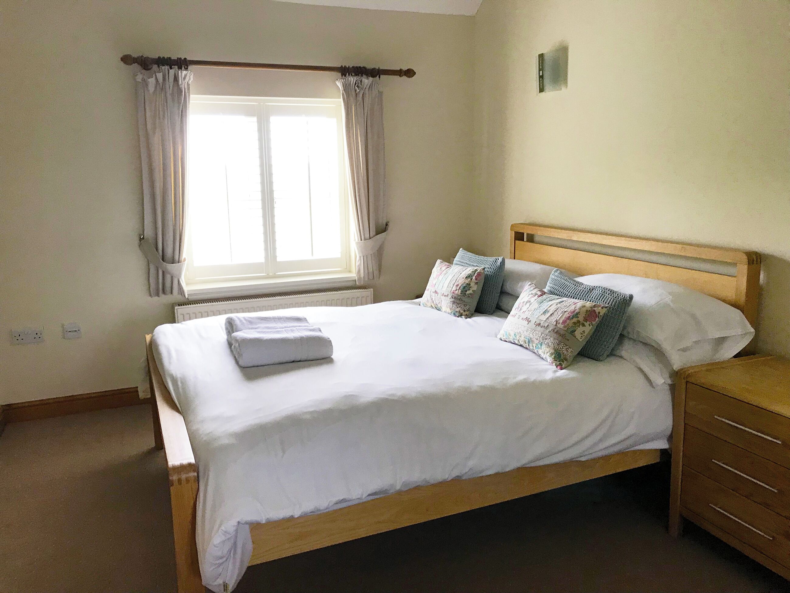A neatly made bed with white linens and decorative pillows, a wooden headboard, and a nightstand with a drawer.