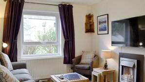 Ein gemütliches Wohnzimmer mit Kamin, einer bequemen Couch, einem kleinen Tisch mit einem Laptop und einem an der Wand befestigten Fernseher.