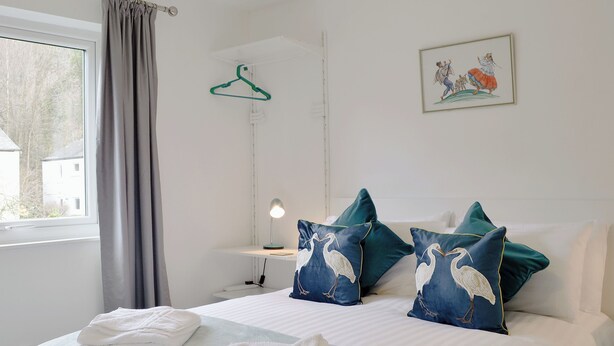 Ein ordentlich bezogenes Bett mit weißen Leinentüchern und zwei türkisfarbenen Kissen mit Vogelmotiven, ein Nachttisch mit Lampe, ein grüner Kleiderhaken und ein gerahmtes Bild an der Wand.