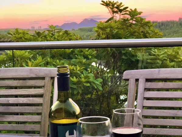 A balcony with a bottle and two glasses, overlooking a landscape with greenery and distant mountains.