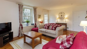 A living room with a sofa, a coffee table, a TV, a clock, and a window with curtains. - 1 bedroom accommodation in Gargrave, near Skipton (Gargrave, near Skipton)