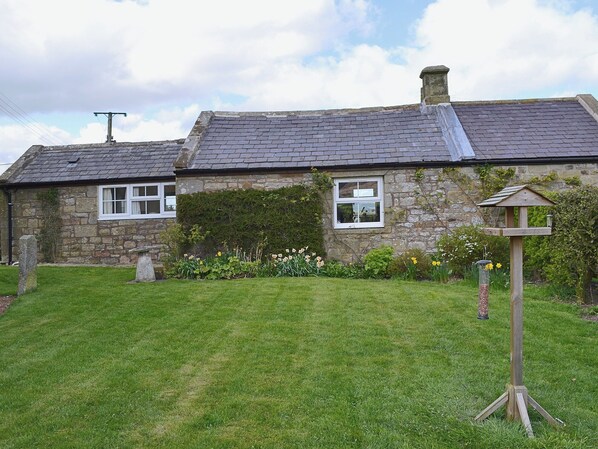 A stone house with a sloped roof, a garden with flowers, and a bird feeder. - 1 bedroom accommodation in Edlingham, near Alnwick (Edlingham, near Alnwick)