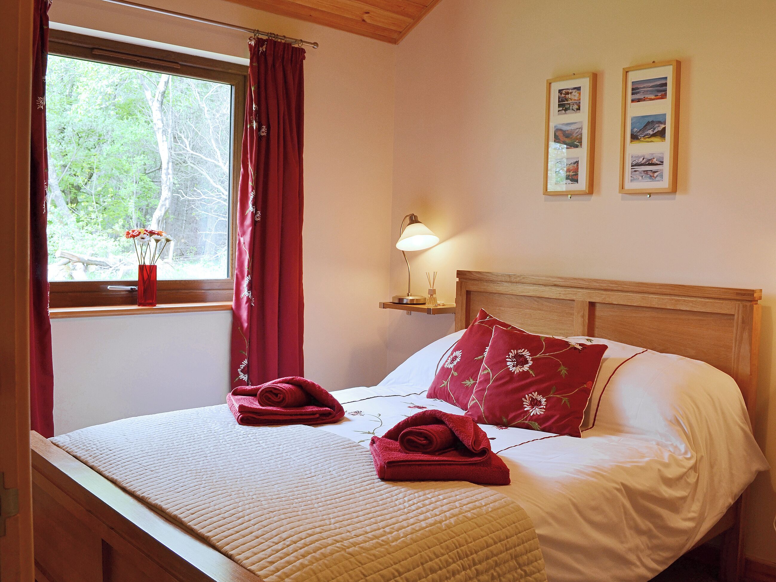 A bed with a wooden headboard, a window with red curtains, a bedside lamp, and two framed pictures on the wall.