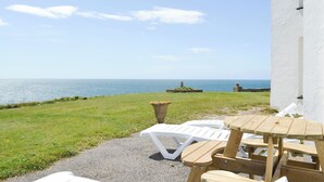 Une terrasse avec des chaises en plastique blanc et des tables en bois, donnant sur une pelouse et la mer.