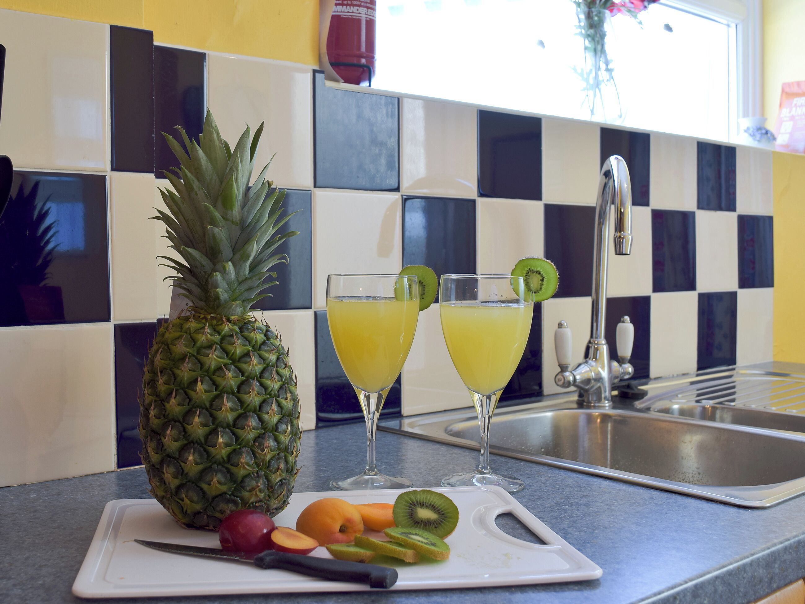 A kitchen counter with a pineapple, sliced fruits, and two glasses of yellow beverage garnished with kiwi slices.