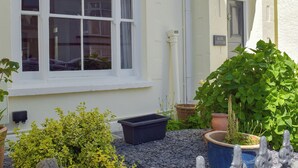 Una cabaña blanca con una ventana, una puerta y un letrero que dice “ROSE COTTAGE”. Hay plantas en macetas y un recipiente negro en el suelo.