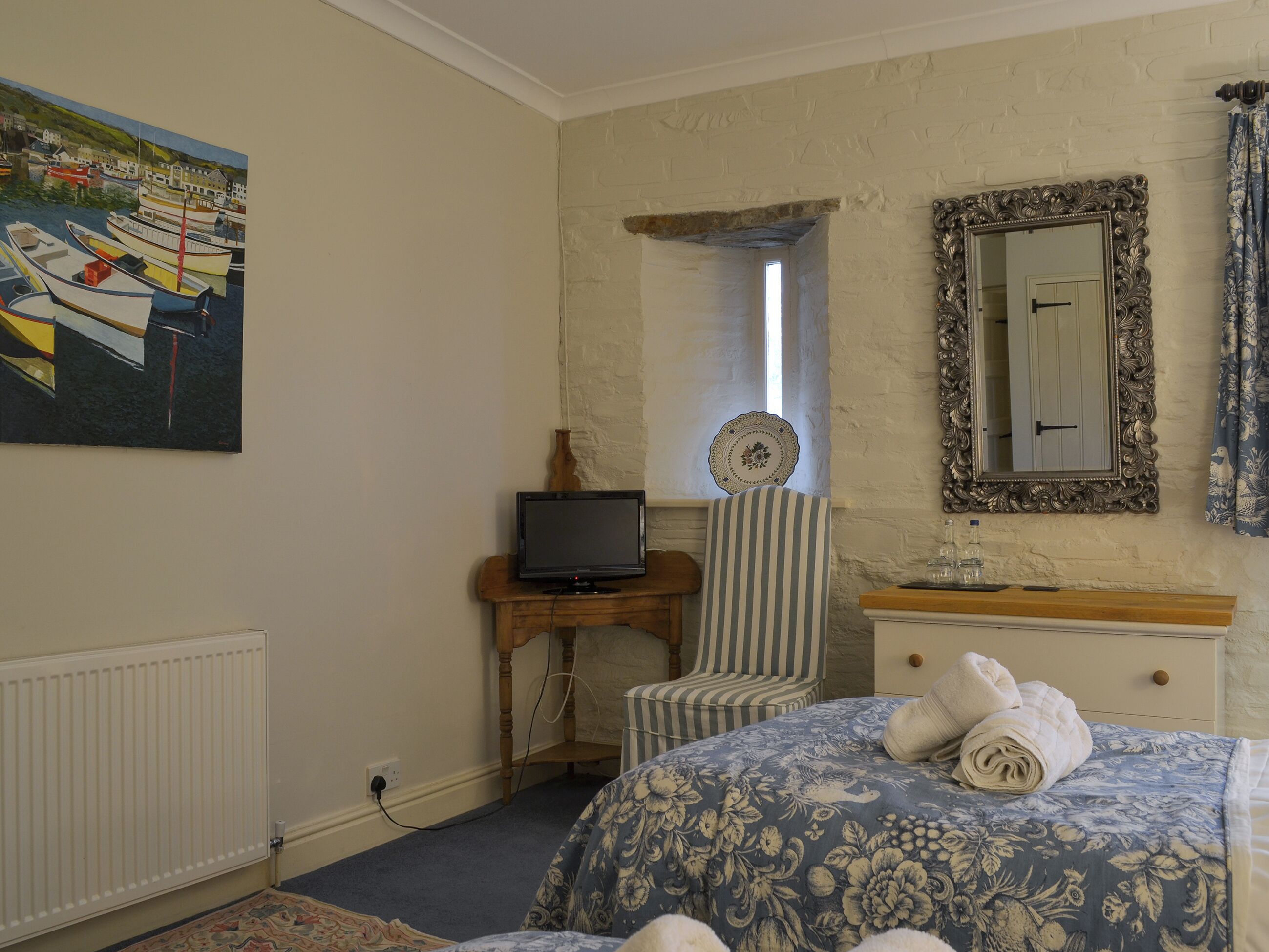 Une chambre à coucher avec un lit, un téléviseur posé sur un meuble en bois, une chaise, une commode, un miroir et un tableau accroché au mur.