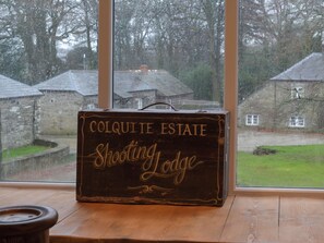 A wooden suitcase with 'Colquitte Estate Shooting Lodge' written on it, placed on a wooden floor near a window overlooking a rural landscape.