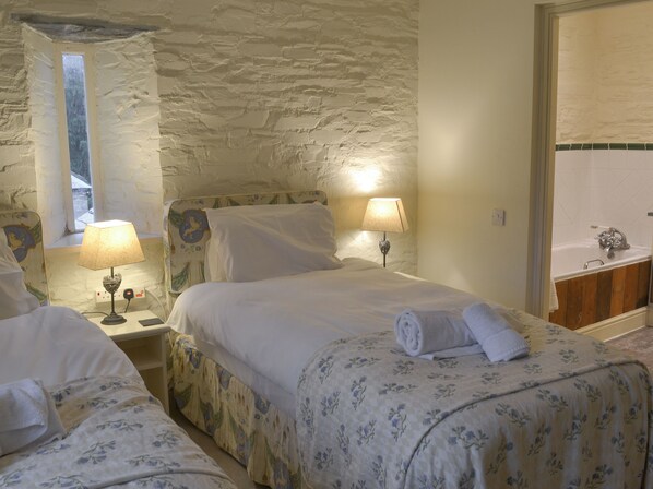 Une chambre double avec deux lits, chacun équipé d’une lampe, d’une fenêtre et donnant sur un mur en pierre.