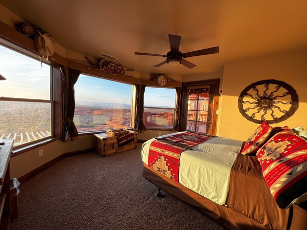 A bedroom with a large bed, a ceiling fan, and a view of a canyon through the windows.