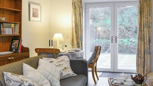 A cozy living room with a sofa, a dining table with chairs, a bookshelf, and a view through a glass door to an outdoor area. - Nettlebush Cottage-UK5629 (Drumelzier, near Peebles)