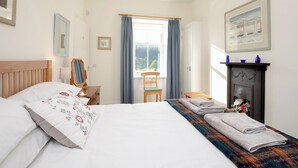 A bedroom with a bed, a chair, a fireplace, and a window with curtains. - Nettlebush Cottage-UK5629 (Drumelzier, near Peebles)