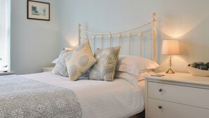 A bedroom with a bed, pillows, a bedside table with a lamp, a nightstand, a drawer, a picture on the wall, and a window with blinds.
