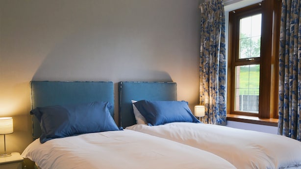 Un lit double avec une literie blanche et bleue, une table de chevet avec une lampe et une fenêtre ornée de rideaux à motifs floraux.