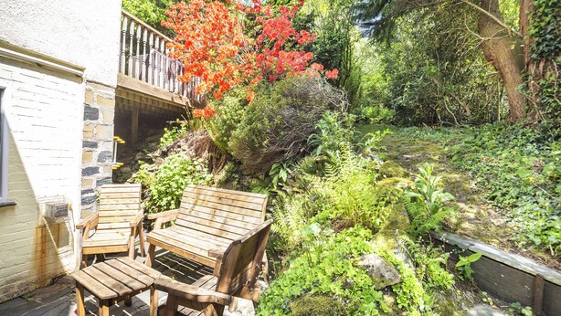 Un banc et une table en bois sont installés sur une terrasse en pierre, avec un jardin en arrière-plan.