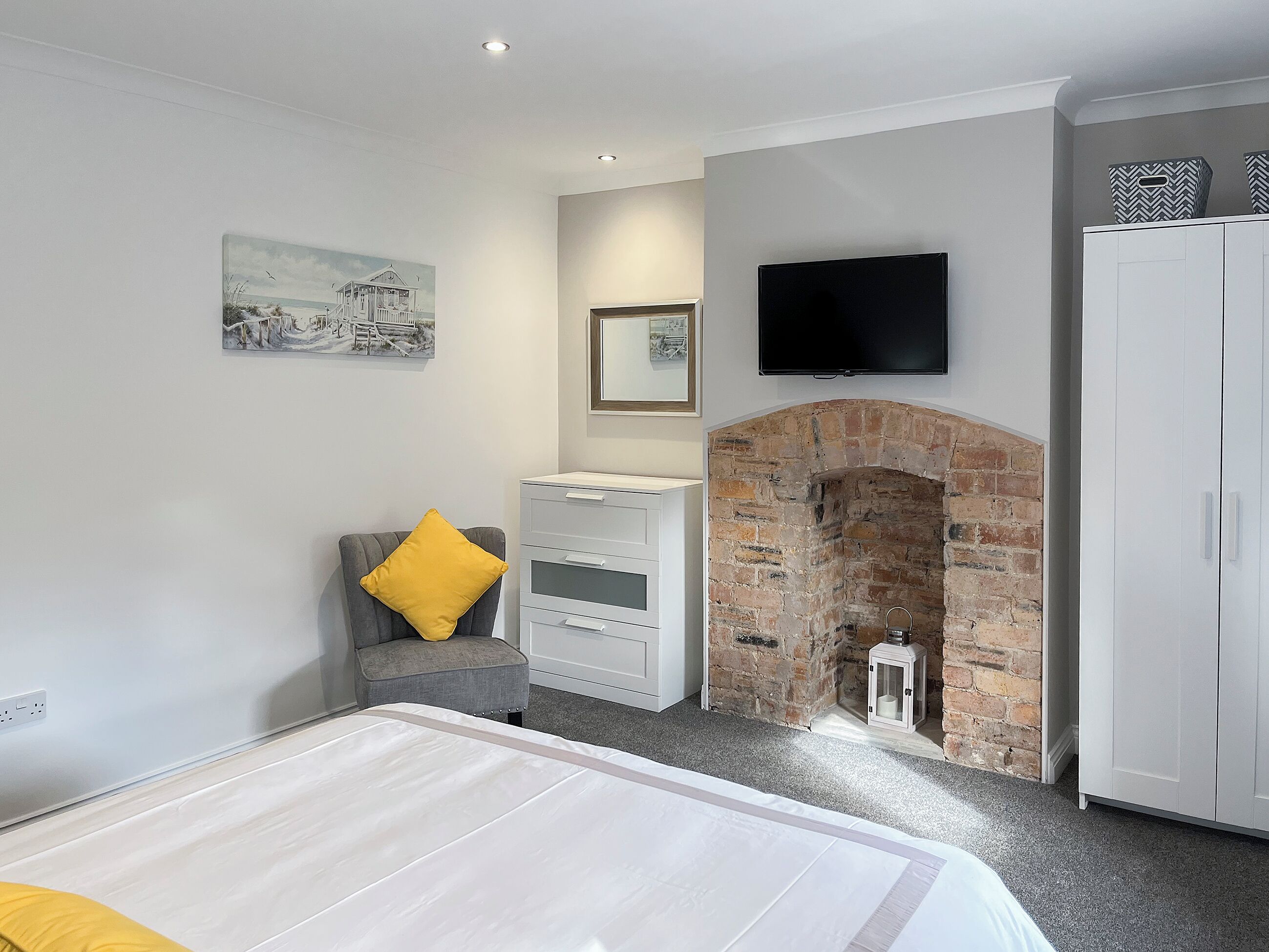 A bedroom with a bed, a gray armchair with a yellow pillow, a white dresser, a brick fireplace, a television mounted above the fireplace, and a wardrobe.