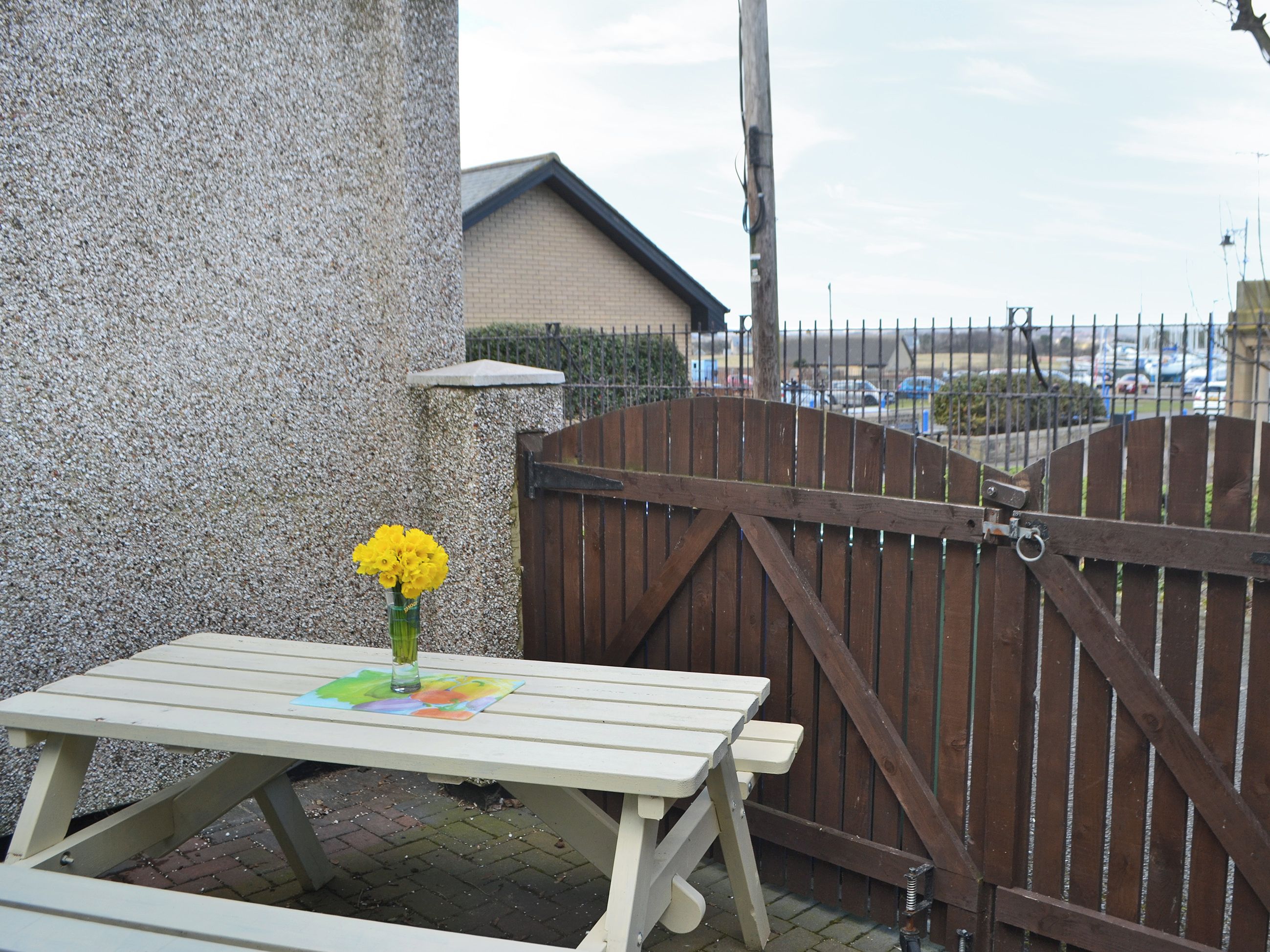 A wooden table with a vase of yellow flowers, a brick wall, and a wooden gate.