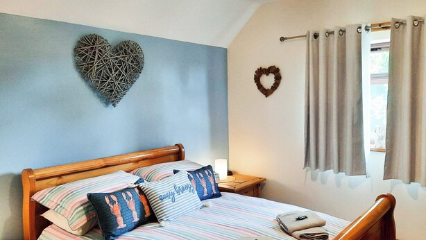 A bed with striped bedding, a heart-shaped wall decoration, a small bedside table with a lamp, and a window with curtains. - 1 bedroom accommodation in Mumby, near Anderby Creek (Mumby, near Anderby Creek)